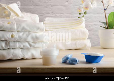 Verschiedene Arten von Waschmitteln auf Holztisch mit frisch gewaschenen gefalteten weißen Kleidern. Vorderansicht. Horizontale Zusammensetzung. Stockfoto