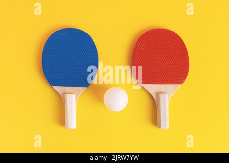 Ein Paar blaue und rote Tischtennisschläger und weiße Kugel auf gelbem Hintergrund. Draufsicht, Studioaufnahme. Tischtennis- oder Tischtennisausrüstung. Stockfoto