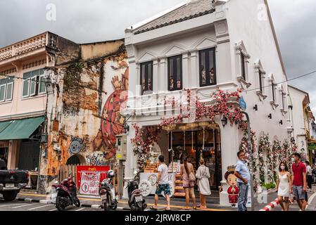 Phuket, Thailand - Juli 2022: Weißes Gebäude mit rosa Blumen an der Thalang Road in der Altstadt von Phuket. Alex Gesicht Wandbild Graffiti. Stockfoto