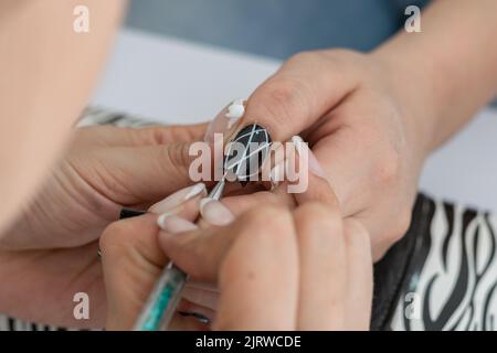 Detailansicht der Hände eines Manikuristen, der lernt, wie man die Nägel einer Frau mit weißen Linien verziert, umreißt mit einem Pinsel. Erstellen eines Maniküre-Designs. conce Stockfoto
