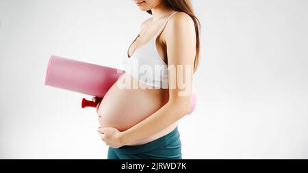 Eine Schwangerin hält eine rosa Yogamatte und streichelt seinen Bauch. Stockfoto