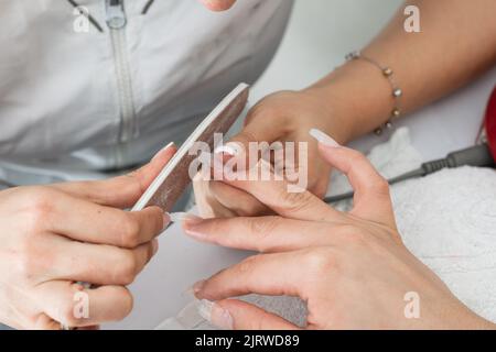 Detailaufnahme einer lateinischen Manikuristin beim Polieren von Polygel-Nägeln mit einer hölzernen Nagelfeile. Konzentriert mit den Fingern ihrer Klientin sehr vorsichtig, um nicht zu sein Stockfoto