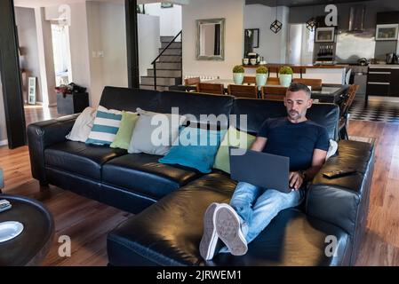 Ein hübscher Mann, der an seinem Laptop arbeitet und zu Hause auf seiner Couch sitzt Stockfoto