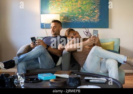 Schwule Paare entspannen sich auf der Couch, während sie Smartphones benutzen Stockfoto