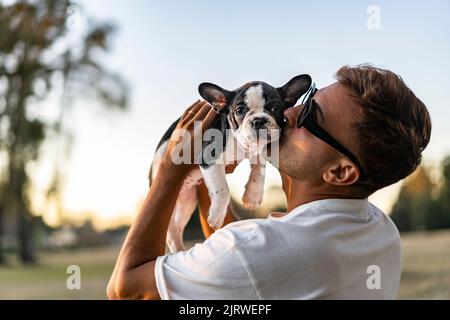 Ein hübscher junger Mann hält sich und küsst eine französische Bulldogge. Stockfoto
