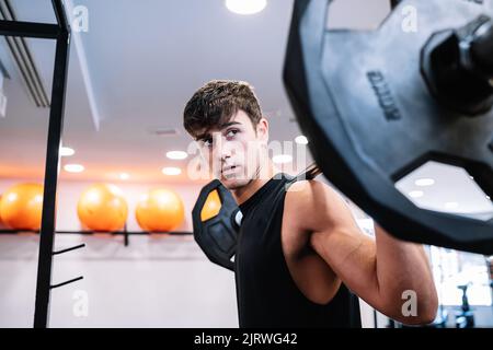 Starker junger Sportler mit Langhantel auf den Schultern, der beim Gewichtheben tagsüber im Fitnessstudio wegschaut Stockfoto