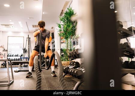 Ganzkörper-entschlossener männlicher Athlet, der in der Nähe der Hantelablage steht und beim Fitnesstraining im Fitnessstudio mit Kampfseilen trainiert Stockfoto