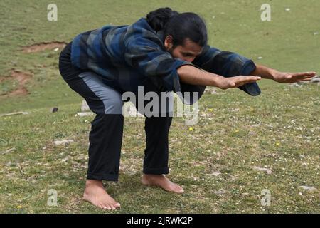 Ein langhaariger junger Inder, der im Freien die Bärenpose (Chair Pose Variation Forward Stretch) praktiziert. Ein junger Mann, der Utkatasana Yoga Pose macht (Variante Vorwärts strecken). Stockfoto