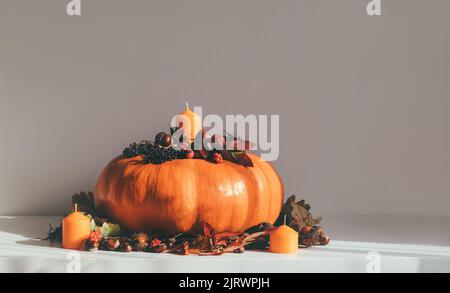 Kürbis mit Kerzen, getrockneten Blättern, Kastanien und Beeren auf einem weißen Tisch. Festliche Herbstdekorationen für Thanksgiving. Stockfoto