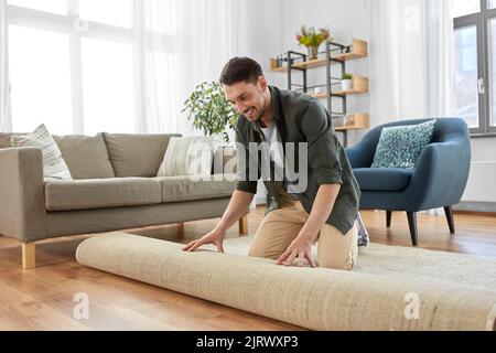 Glücklich lächelnder junger Mann, der sich zu Hause den Teppich ausbreitet Stockfoto