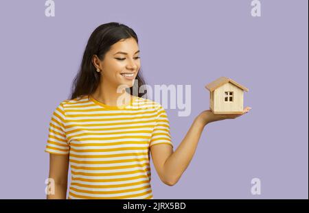 Frau, die davon träumt, ein Haus zu besitzen, mit einem Modell eines isolierten Hauses auf pastellviolettem Hintergrund. Stockfoto