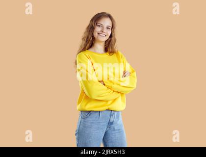 Junge lächelnde Frau in gelbem Sweatshirt und Jeans posiert auf beigem Hintergrund und blickt auf die Kamera. Stockfoto