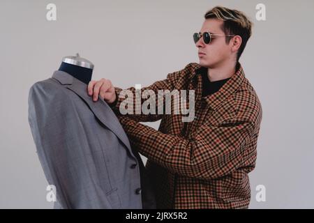 Ein junger Modedesigner bei der Arbeit in einem grauen Anzug. Kaukasische blonde Modedesignerin in einem gelben Anzug und einer schwarzen Brille, die an einer Schaufensterpuppe arbeitet. Hochwertige Fotos Stockfoto