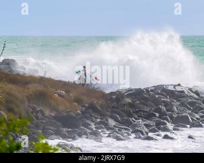 Raglan, Neuseeland - 7 2008. Juni; junger Surfer klettert über felsige Küste, um zu surfen, während riesige Wellen um ihn herum hereinstürzen. Stockfoto