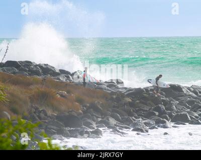 Raglan, Neuseeland -Juni 7 2008 Junge männliche Surfer klettern über felsige Küste, um zu surfen, während riesige Wellen um ihn herum hereinstürzen. Stockfoto