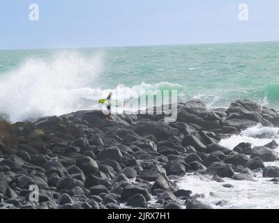 Raglan, Neuseeland - 7 2008. Juni; junger Surfer klettert über felsige Küste, um zu surfen, während riesige Wellen um ihn herum hereinstürzen. Stockfoto