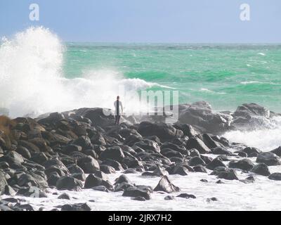 Raglan, Neuseeland - 7 2008. Juni; junger Surfer klettert über felsige Küste, um zu surfen, während riesige Wellen um ihn herum hereinstürzen. Stockfoto