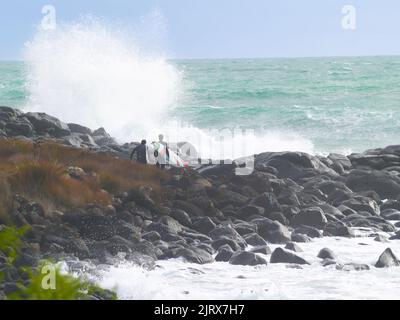 Raglan, Neuseeland -Juni 7 2008 junger Surfer klettert über felsige Küstenlinie, um zu surfen, während riesige Wellen um ihn hereinstürzen. Stockfoto