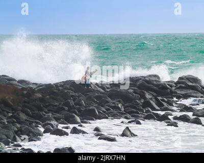 Raglan, Neuseeland - 7 2008. Juni; junger Surfer klettert über felsige Küste, um zu surfen, während riesige Wellen um ihn herum hereinstürzen. Stockfoto