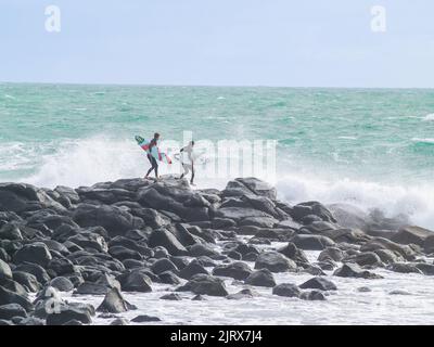 Raglan, Neuseeland -Juni 7 2008 Junge männliche Surfer klettern über felsige Küste, um zu surfen, während riesige Wellen um ihn herum hereinstürzen. Stockfoto