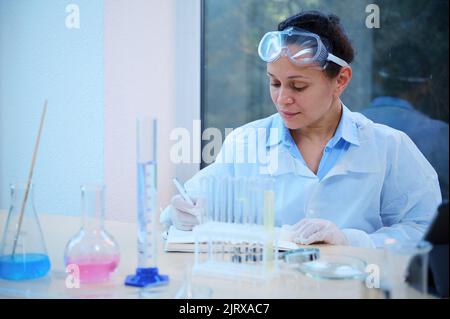 Der Chemiker und Pharmakologe schreibt die Ergebnisse von Experimenten in einem pharmakologischen Labor auf einen Notizblock. Stockfoto