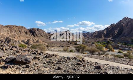 Landschaftsansicht von Wadi Shawka trockenem Flussbett mit Emirates Adventures Lagergebäuden, felsigen Hajar Mountains im Hintergrund, Vereinigte Arabische Emirate. Stockfoto