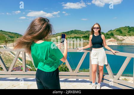 Ein junges Mädchen macht mit einem Handy ein Foto von ihrer Freundin vor dem See. Das Konzept der Einflussnehmer auf Reisen. Berg im Hintergrund Stockfoto