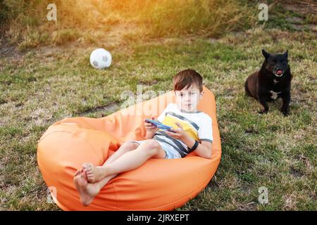 Happy Kid Boy Spiel auf dem Smartphone im Park im Freien, Kind mit dem Smartphone auf weichen orangefarbenen Stuhl zu Hause Garten nach aktiven Spielen mit Ball und Hund, Hinterhof, Sonnenlicht, Handy sucht Stockfoto