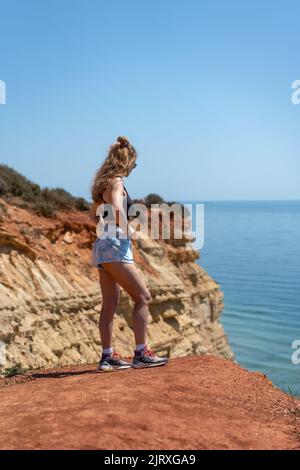 Eine junge, blonde Frau, die auf einer Klippe posiert und unter der Sonne über den Ozean blickt Stockfoto