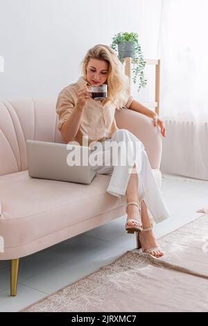 Eine blonde Geschäftsfrau, die auf einem Laptop arbeitet, sitzt auf dem Sofa mit einem Kaffeetasse Stockfoto