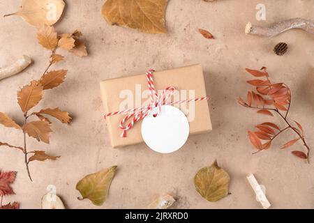 Herbsthintergrund: Gefallene Blätter, trockene Pflanzen mit handgefertigten Geschenken, eingewickelt in Bastelpapier und Seil mit Geschenkanhänger-Mockup auf natürlichem beigen Hintergrund. Tha Stockfoto