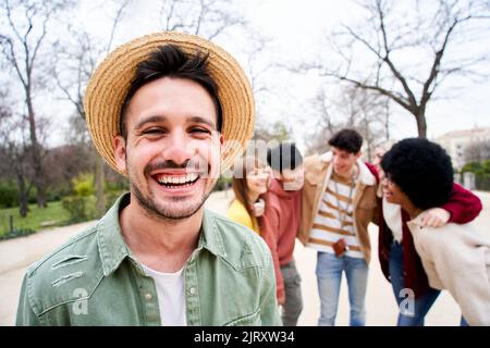 Junger lächelnder Kerl, der mit einer Gruppe von Freunden die Kamera im Freien anschaut. Glückliche Menschen gemeinsam Spaß haben Konzentrieren Sie sich auf einen hübschen jungen Mann. Universität Stockfoto