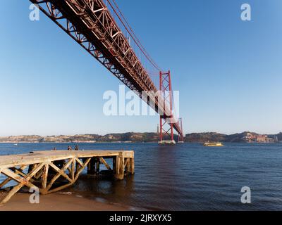 Ponte 25 de Abril (Brücke vom 25. April) über den Fluss Tejo in Lissabon, Portugal. Anlegestelle und Strand links. Stockfoto