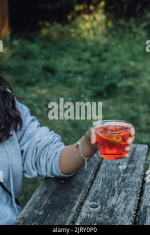 Frau mit Aperol-Spritz-Cocktail mit grüner Olivgarnitur, im Sommer draußen am Picknicktisch Stockfoto