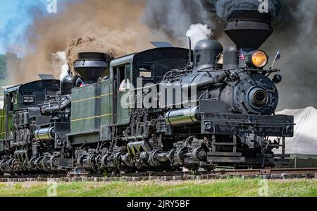 Blick auf zwei Shay Dampfmaschinen, die sich auf eine Dampfparade mit den beiden miteinander verbundenen Motoren aufmachen, die an einem sonnigen Sommertag schwarzen Rauch und weißen Dampf blasen Stockfoto