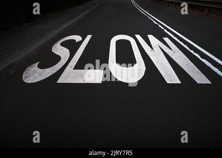 SLOW Road Schild auf Bitumen Asphalt Straße in weißer Farbe lackiert. Niedrige, weit abgewinkelte Perspektive. Verwitterung und Verschleiß sind an der Oberfläche sichtbar Stockfoto