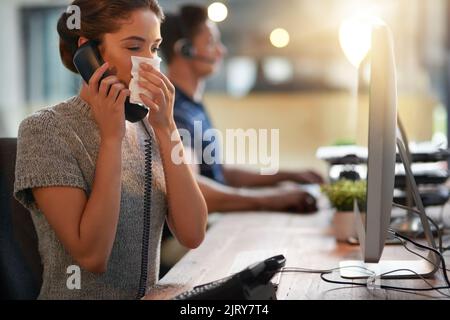 Es ist schwer, Arbeit zu erledigen, wenn Sie sich unwohl fühlen. Eine junge Geschäftsfrau bläst ihre Nase, während sie in einem Büro am Telefon telefoniert. Stockfoto