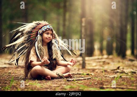 Ihr Erbe von einem frühen Alter an zu verstehen. Ganzkörperaufnahme eines kleinen Mädchens, das meditiert, während es im Wald Dressup spielt. Stockfoto