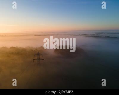 Luftaufnahme eines nebligen Sonnenaufgangs mit Stromleitungen, die wie eine Reihe von Soldaten im Nebel verschwinden Stockfoto