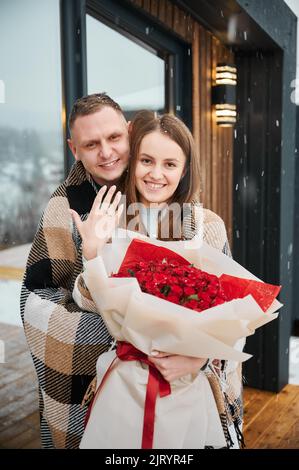 Glückliche Frau, die einen Strauß Rosen hält und Hand mit Verlobungsring zeigt, während der Mann den Verlobten von hinten umarmt. Porträt eines verlobt verliebten Paares, das am Wintertag vor dem Haus steht. Stockfoto