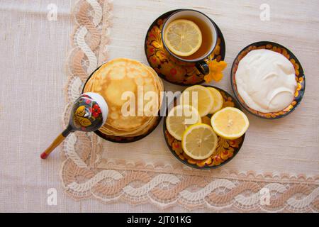 Tee in einer bemalten Tasse, auf einer Untertasse in Scheiben geschnitten Zitrone, Pfannkuchen, saure Creme und einem Holzlöffel zum Überlegen. Tee-Party im rustikalen Stil Stockfoto