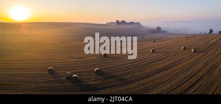 Leuchtend gelbe aufgehende Sonne über goldenen Feldern und Heuballen mit Nebel und Baumsihlouetten im Hintergrund. Landschaft von Roztocze Polen. Horizontale Aufnahme. Hochwertige Fotos Stockfoto