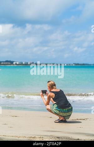 Frau fotografiert am Strand mit dem Smartphone gegen blaues Wasser und Wolken Stockfoto