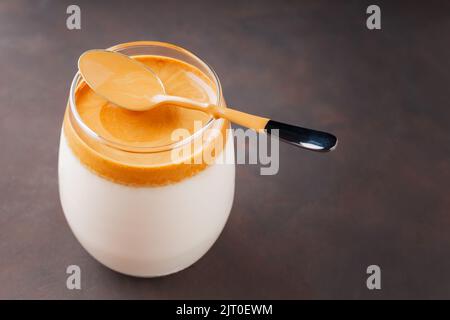 Dalgona Kaffee mit einem Löffel auf einem dunkelbraunen Hintergrund. Eisgekühltes, flauschiges, Schlagtrend-Getränk mit Kaffeeschaum und Milch. Koreanischer trendiger Kaffeedrink Stockfoto