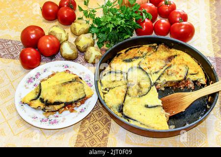 Der heiße, fertige Moussaka in der Form zum Backen wird von einem Holzspatel unter Gemüse und würzigen Kräutern in Stücke geschnitten. Ein Stück des Tellers wird auf einem serviert Stockfoto