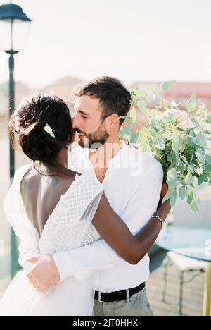 Der Bräutigam umarmt die Braut mit einem Blumenstrauß auf dem Dach des Gebäudes Stockfoto