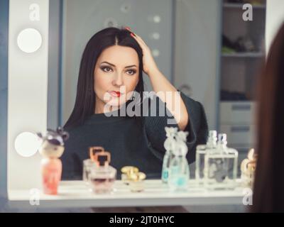 Nachdenkliche junge Frau in warmer grüner Kleidung, die Spiegelung im Spiegel betrachtet und ihr Haar mit der Hand kämmt. Stockfoto