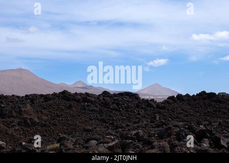 Lavaboden von brauner bis schwarzer Farbe. Berge im Hintergrund. Blauer Himmel mit weißen Wolken im frühen Frühjahr. Lanzarote, Las Palmas, Kanarische Inseln Stockfoto