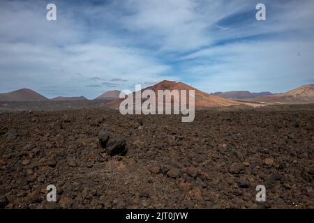 Lavaboden von brauner bis schwarzer Farbe. Berge im Hintergrund. Blauer Himmel mit weißen Wolken im frühen Frühjahr. Lanzarote, Las Palmas, Kanarische Inseln Stockfoto