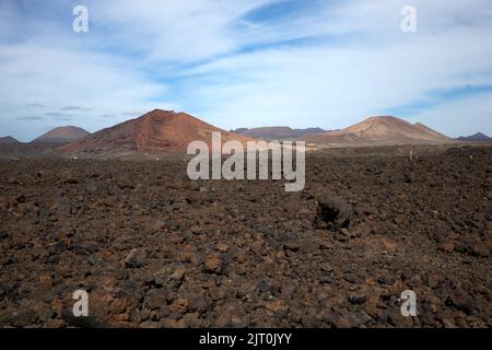 Lavaboden von brauner bis schwarzer Farbe. Berge im Hintergrund. Blauer Himmel mit weißen Wolken im frühen Frühjahr. Lanzarote, Las Palmas, Kanarische Inseln Stockfoto